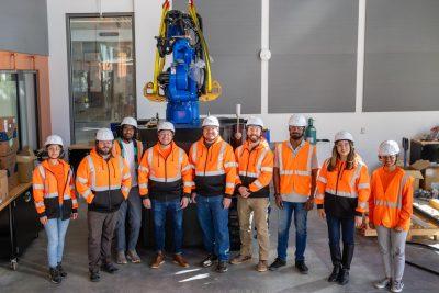 A group of people in reflective orange shirts and hardhats stand in front of large 3D printer with robotic arm.